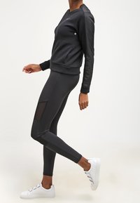 Sudadera negra con tela texturizada, cuello redondo y mangas largas; combinada con leggings negros ajustados que tienen paneles sheer. Zapatillas blancas.