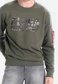Uomo che indossa un felpa oliva di Alpha Industries con logo camouflage, etichetta rossa sulla manica e jeans grigi sbiaditi, in piedi con le mani nelle tasche.