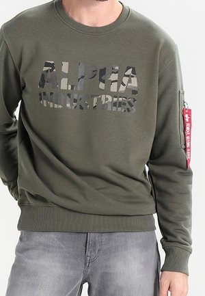Hombre vestido con un sudadera oliva de Alpha Industries con logo de camuflaje, etiqueta roja en la manga y jeans grises deslavados, de pie con las manos en los bolsillos.