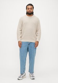 Maglione beige chiaro lavorato a maglia con motivo strutturato, abbinato a jeans di denim blu larghi e scarpe sportive grigie e bianche.