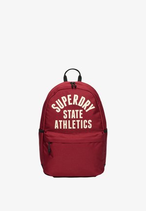 Sac à dos rouge avec fermetures éclair et poignée noires, affichant un grand texte blanc "SUPERDRY STATE ATHLETICS" sur la poche avant.