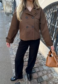 Brown double-breasted jacket in textured fabric, paired with black wide-leg pants and shiny black loafers. Brown leather handbag accent.