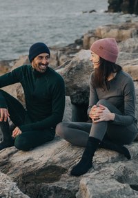 Two individuals sit on rocky terrain. One wears a dark green zip-up top, matching pants, and a navy beanie. The other is in a grey top, leggings, and a pink beanie.