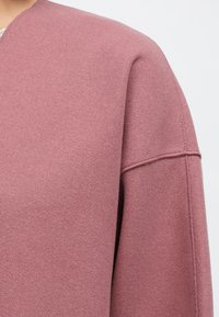 Rosa, strukturerat tyg på ett plagg med rund halsringning, sänkta axlar och rena sömnadsdetaljer. Mjuk, varm känsla.