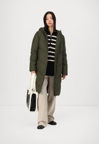 Abrigo de plumas verde oliva con capucha, cierre de cremallera y diseño acolchado, combinado con una camiseta a rayas en blanco y negro, pantalones anchos beige y un bolso negro con borde de piel.