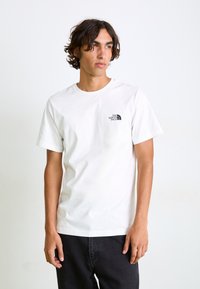 Hvid bomulds t-shirt med rund halsudskæring og korte ærmer, med et lille sort logo på venstre bryst. Båret med mørke jeans.