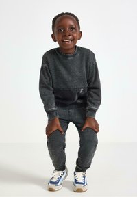 Felpa nera con tasca frontale, jeans in denim scuro e sneakers blu con accenti bianchi. Soggetto sorridente, capelli corti e ricci.