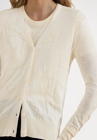 Béžový sveter s jemnou pleteninou, výstrihom do V, zapínaním na gombíky a zložitými vzormi tkanými do látky.