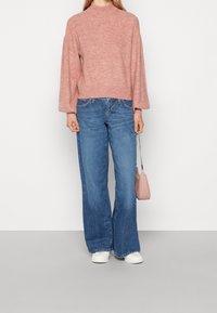 Rosa stickad tröja med hög hals, kombinerad med ljusblå vida jeans. Vita sneakers och en liten rosa handväska fulländar looken.