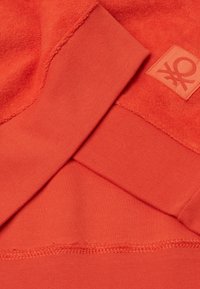 Felpa arancione con tessuto che presenta una parte superiore in pile testurizzato e una parte inferiore in maglia liscia. Include un'etichette con logo e cuciture visibili.