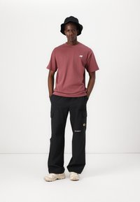 Bordeauxfarbenes T-Shirt mit kurzen Ärmeln und einem Logo, kombiniert mit schwarzen Cargo-Hosen mit Taschen und einem gelben Emblem, getragen mit beigen Sneakers.