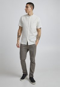 Chemise en lin blanche à manches courtes avec un col à bande et une poche avant, associée à un pantalon chino gris clair et des baskets noires.