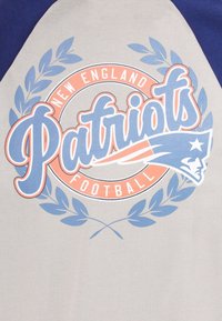 Grå t-shirt med blå ärmar som har en stiliserad "Patriots"-logotyp, lagerdesign och "New England Football" i en orange cirkel.