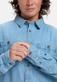 Lichtblauw denim overhemd met knoopsluiting bij de manchetten, borstzakken en koperen knopen. Heeft een klassieke kraag en een gestructureerde stof.