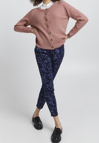 Cardigan rose tricoté avec des boutons, porté sur une chemise blanche à col. Pantalon slim à motifs bleus et noirs et mocassins noirs.