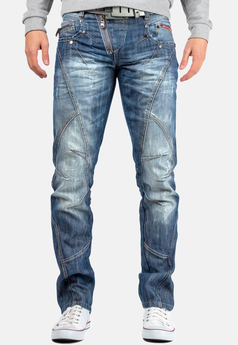 Jean en denim bleu avec une coupe slim, finition délavée et détails de couture asymétriques. Comprend plusieurs poches et une fermeture éclair.