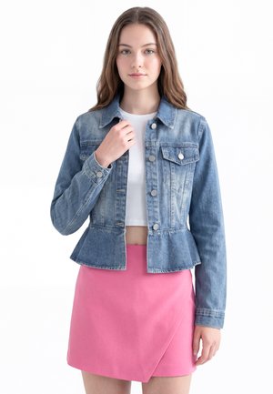 Ung kvinde med langt hår iført en blå denimjakke over en hvid crop top og en pink nederdel, står mod en hvid baggrund.
