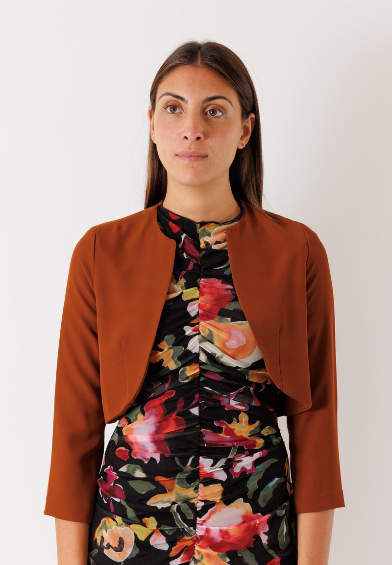 Giacca marrone cropped con maniche a tre quarti sopra un vestito nero aderente con un vivace motivo floreale e dettagli di arricciatura.