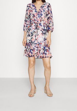 Femme portant une robe fleurie mi-longue avec un décolleté en V, des manches 3/4 bouffantes, un ourlet volanté et des sandales à talons blocs beige.