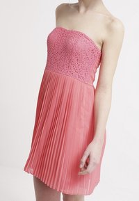 Šaty bez ramínek v korálově růžové barvě; korzet zdobí spletitá krajka, zatímco sukně je vyrobena z lehkého materiálu a jemně splývá.