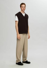 Brown knitted sleeveless vest over a white t-shirt, paired with light beige trousers and black shoes. Simple, relaxed fit.