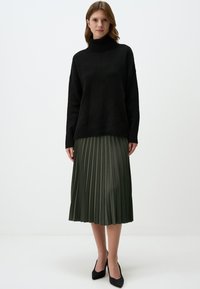 Schwarzer übergroßer Rollkragenpullover mit gerippten Bündchen, kombiniert mit einem plissierten olivgrünen Midirock. Das Model trägt schwarze spitze Absatzschuhe.