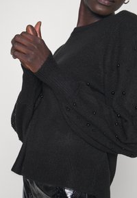 Maglione nero lavorato a maglia con maniche puff testurizzate decorate con perline nere, dotato di scollatura rotonda e polsini a coste.