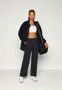 Kvinne i svart zip-up jakke, hvit sports-bh, svarte vide bukser, hvite joggesko, bærende svart skulderveske, stående mot en enkel bakgrunn.