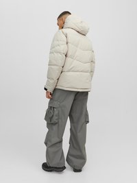 Beige Daunenjacke mit Kapuze, featuring gesteppten Abschnitten. Kombiniert mit grauen Cargo-Hosen, die seitliche Taschen und eine lockere Passform zeigen.