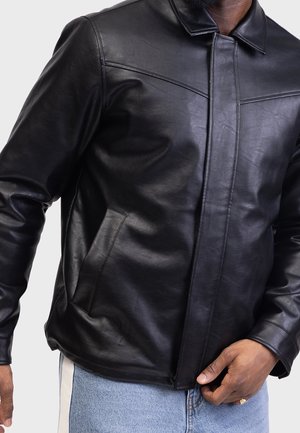 Veste en cuir noire avec poches avant portée par une personne en jean bleu clair, main ajustant la veste près de la taille, fond clair uni.