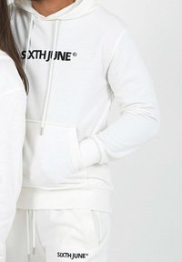 Sudadera blanca con bolsillo frontal y cordón ajustable. Presenta el logo "SIXTHJUNE" en negro en el pecho. Textura suave.