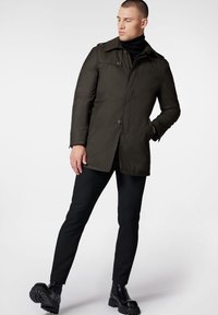 Oliwkowy płaszcz z zapięciem na guziki, dużym kołnierzem i bocznymi kieszeniami, noszony na czarnym golfie i zestawiony z dopasowanymi czarnymi spodniami.