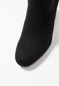 Primer plano de la parte delantera y lateral de una bota de tobillo de ante negro sobre un fondo blanco.