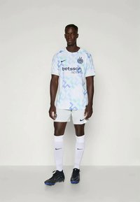 Muški nogometaš stoji, obučen u bijelu i svijetloplavu majicu sa uzorkom Inter Milana, bijele kratke hlače, bijele čarape i crne kopačke.