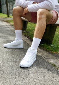 Hombre con zapatillas blancas, calcetines blancos, pantalones cortos rosas y una sudadera ligera sentado en un banco del parque con las manos apoyadas sobre las rodillas.