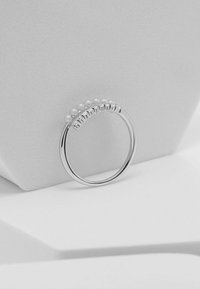 Anello d'argento con una fila di quattro piccole perle e pietre trasparenti alternate, montato su una fascia lucida, su uno sfondo chiaro e strutturato.