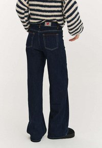 Tmavě modré džíny s širokými nohavicemi, obsahující pět kapes, prošité detaily a vzorovanou štítek na pásku. Hladká textura tkaniny.