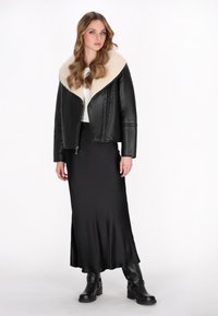 Schwarze Lederjacke mit einem breiten Kragen aus cremefarbiger Schafswolle, kombiniert mit einem langen schwarzen Satinkleid und schwarzen Knöchelstiefeln mit Schnallen.