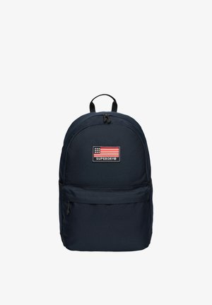 Marineblauwe rugzak met een voorvak met ritssluiting, bovenste handvat en een patch met een Amerikaanse vlag en het "SUPERDRY"-logo.