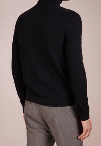 Pull en maille noir à col roulé avec col côtelé et détails texturés sur les épaules, associé à un pantalon à carreaux avec des accents orange.