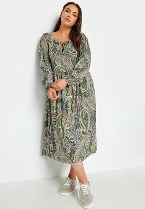 Abito a maniche lunghe verde con motivo paisley, vita regolata, scollatura rotonda e dettaglio a chiave, abbinato a sneakers grigie.