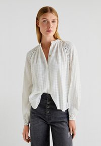 Blusa bianca a maniche lunghe con una trama textured, caratterizzata da un collo rotondo, bottoni frontali e dettagli arricciati sulle spalle e sui polsini.