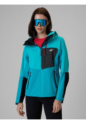 Kobieta ubrana w niebieską i czarną kurtkę outdoor z kapturem oraz duże, refleksyjne niebieskie sportowe okulary przeciwsłoneczne na jednolitym szarym tle.