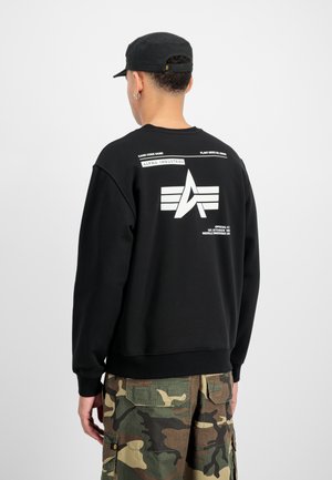 Felpa nera con un logo grafico bianco sul retro, vestibilità rilassata e polsini a coste. Abbinata a pantaloni cargo mimetici.
