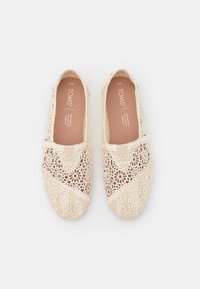 Zapatillas de crochet beige tipo slip-on con patrón floral, que presentan una superficie texturizada y un panel elástico para facilitar su uso. Ideales para entornos casuales.