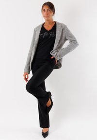 Blazer grigio testurizzato sopra una maglietta nera con scollo a V e testo stampato, abbinato a pantaloni neri aderenti e tacchi a punta neri.