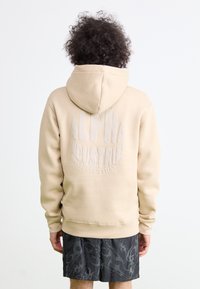 Felpa con cappuccio beige con un grande logo testurizzato "ALPHA INDUSTRIES" sulla schiena. Polsini e orlo a costine. Indossata con pantaloni corti neri a fantasia.