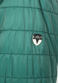 La doudoune verte présente un design matelassé avec des lignes verticales, une texture brillante et un écusson logo CMP en forme de bouclier.