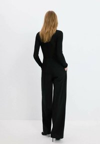 Mujer con cabello largo, vestida con una blusa negra de manga larga y pantalones negros de pierna ancha, de pie con las manos en los bolsillos, mirando hacia atrás.