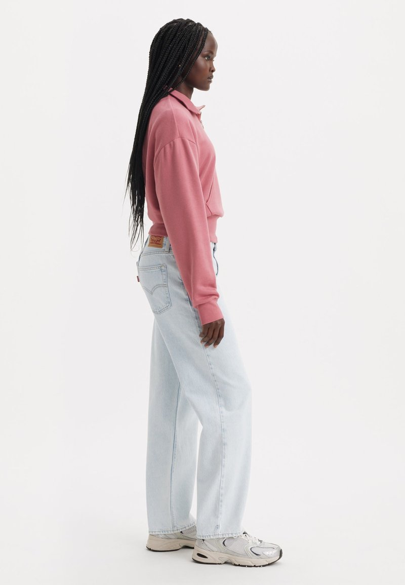 Pullover rosa con colletto, jeans a gamba dritta blu chiaro e sneakers grigie; lunghi capelli intrecciati scendono. Design minimalista.